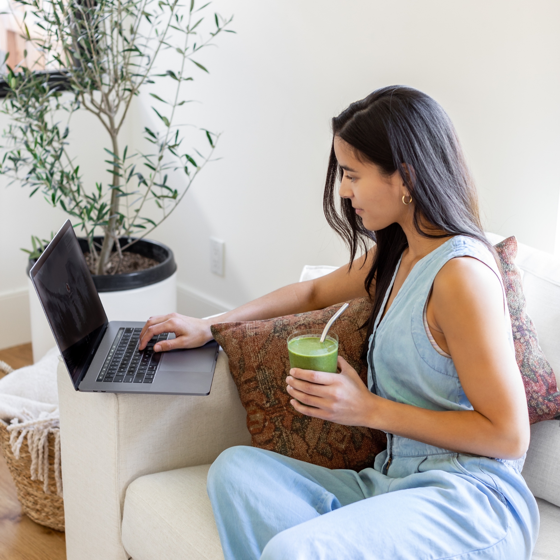 Person sitting on couch holding green smoothie and working on laptop computer.
