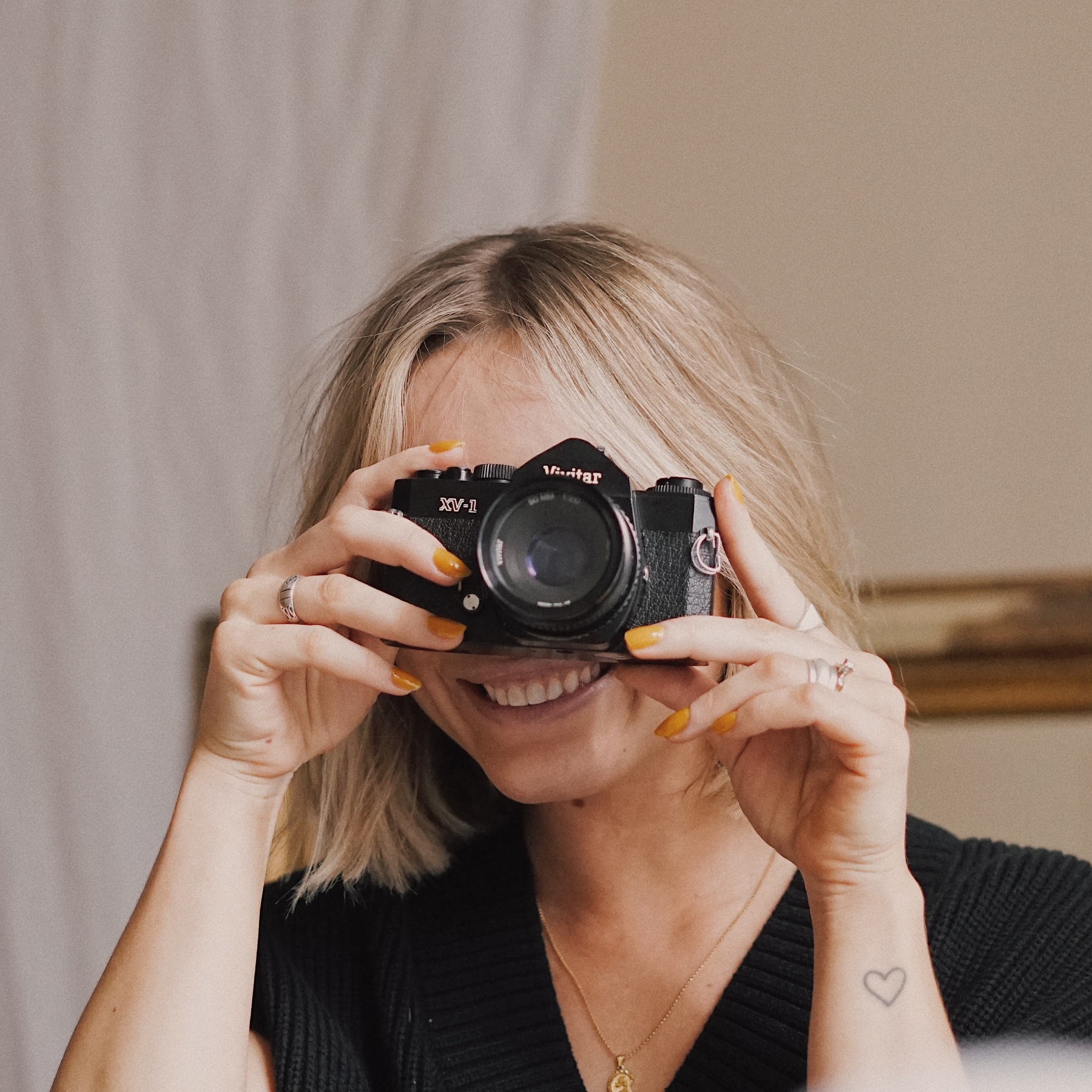 Detail of person with blonde hair, sweater, and heart tattoo holding a black film camera.