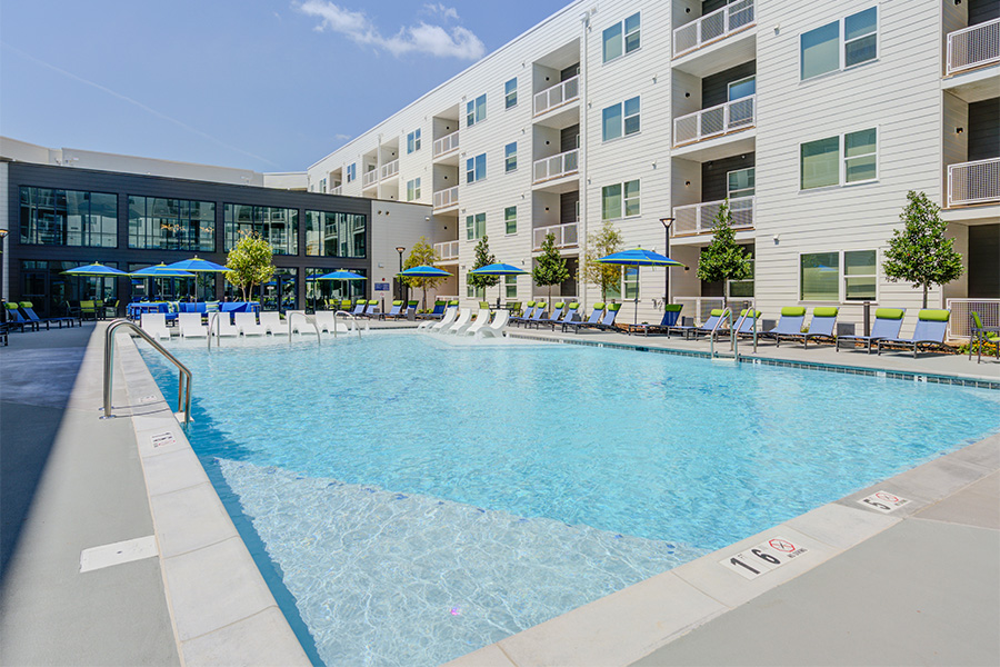 Pet-Friendly Apartments in Huntsville, AL - Constellation - Resort-Style Pool and Tanning Ledge with Lounge Seats, Umbrellas, and View of Apartment Building.